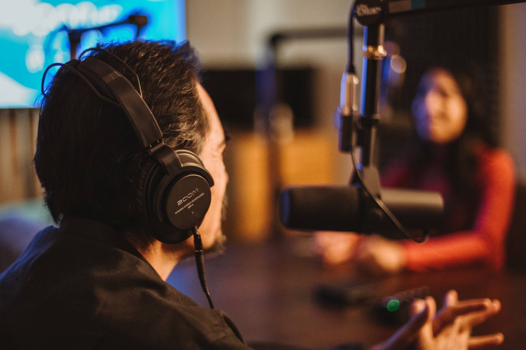 A podcast host with headphones at the microphone, talking with a guest in the studio